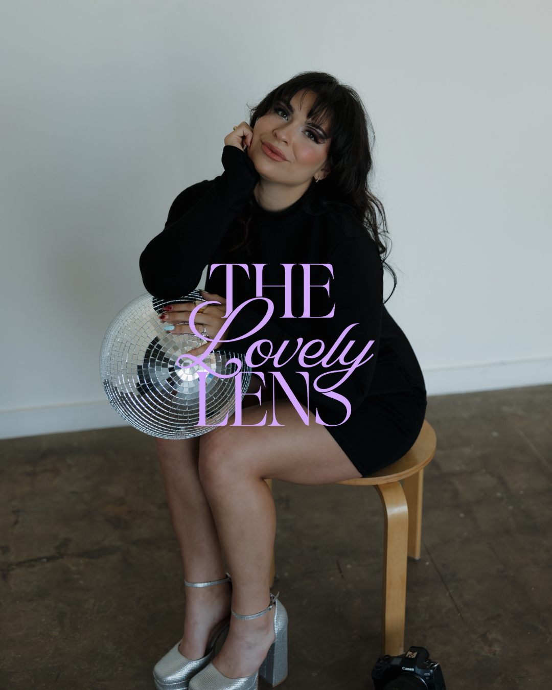 A woman in a black dress sits on a stool holding a disco ball, with "The Lovely Lens" text overlaid on the image.