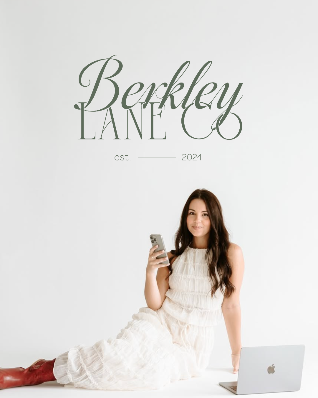 A woman in a white dress sits on the floor with a smartphone and laptop, in front of a wall sign that reads "Berkley Lane Co est. 2024.
