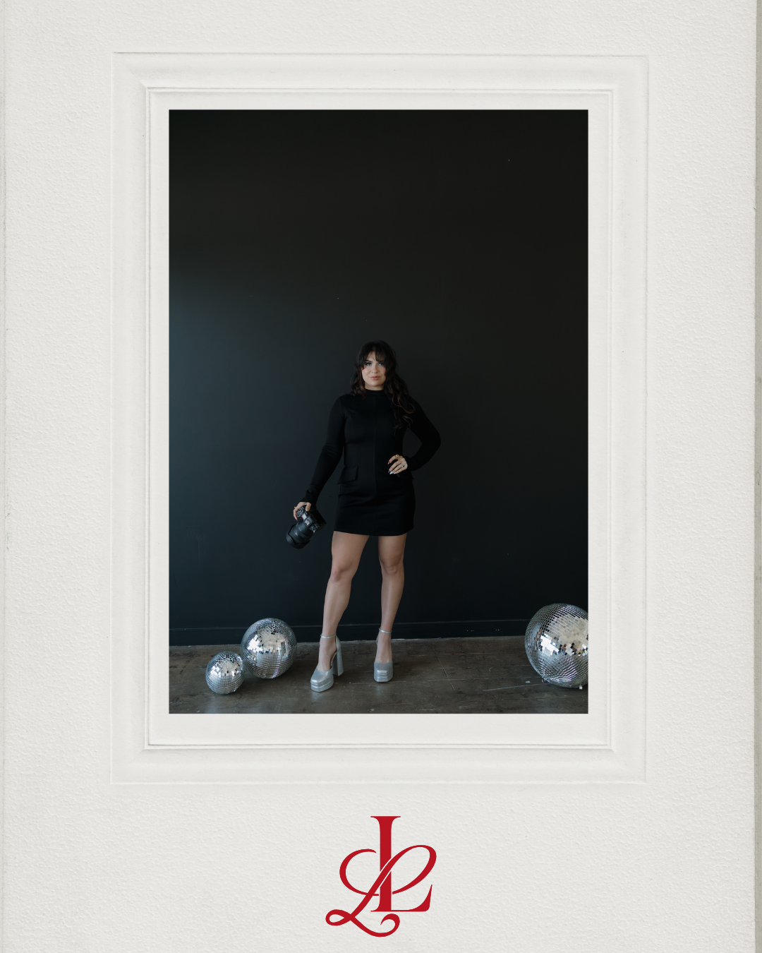 A woman in a black dress and high heels stands against a dark wall, holding a camera, with two disco balls on the floor nearby. The image is framed in white with a red "L" logo at the bottom.